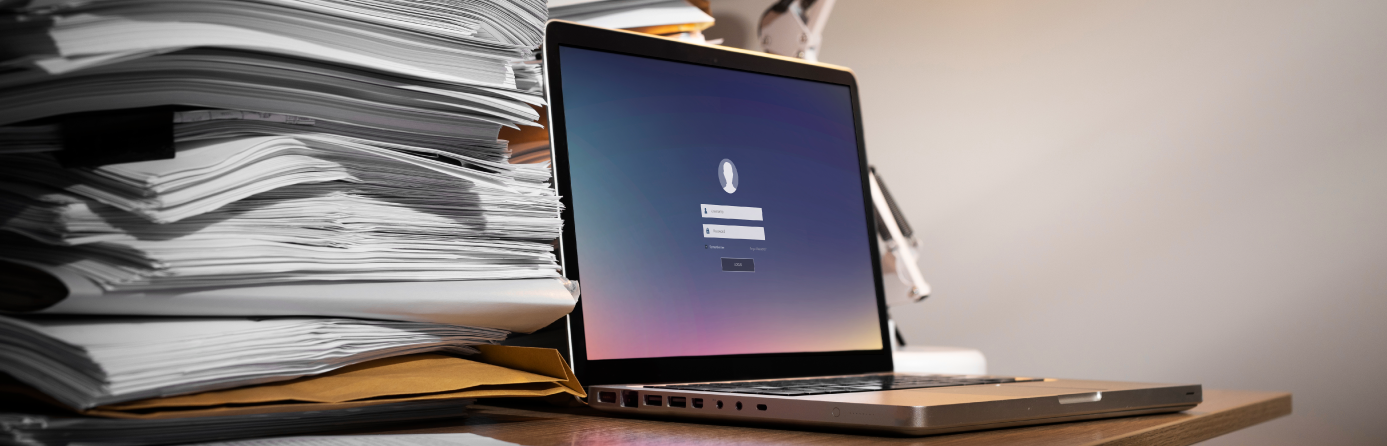 Laptop beside stacks of paper documents on a desk.